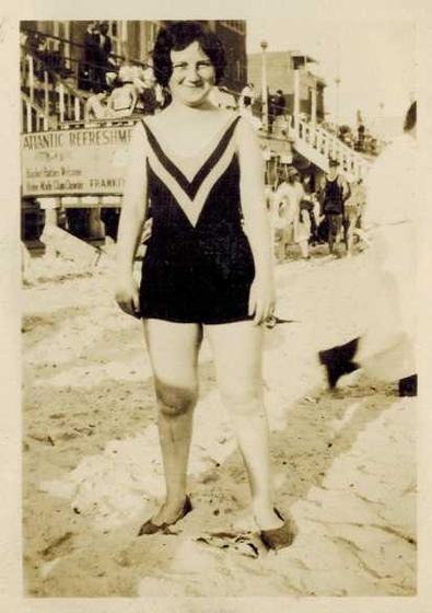 Believed to be Mae on the beach, a refreshment stand behind her.