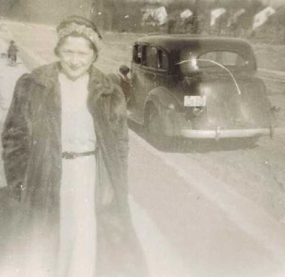 Believed to be Mae in a fur coat beside an old car.