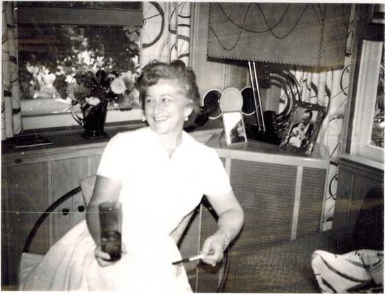 Mae holding a glass, a window with flowers behind her.