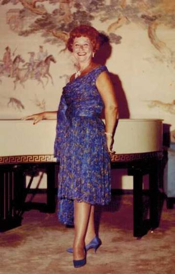 Mae in a blue dress by a white piano and a painted mural.