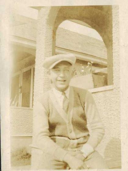 A man in a flat cap and sweater vest, sitting by a stucco archway.