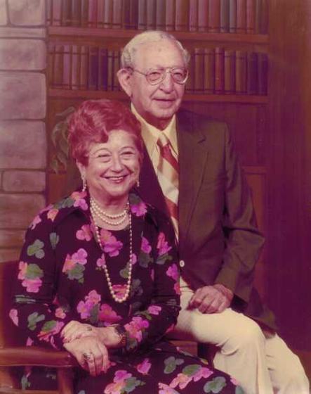Harold and Mae in a formal portrait in front of a bookshelf and a stone wall. Mae wears a floral top and pearls; Harold wears a green jacket and a striped tie.