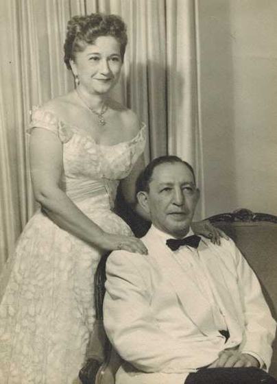 A formal studio portrait: Mae standing in a lace gown, Harold seated in a white dinner jacket and bow tie.