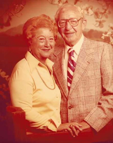 Harold and Mae in a portrait against a mountain backdrop. Mae wears a yellow blouse and a gold chain; Harold wears a plaid jacket and a striped tie.
