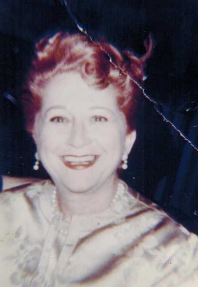 Mae, smiling, in a satin top and pearls. The print has a crease across it.