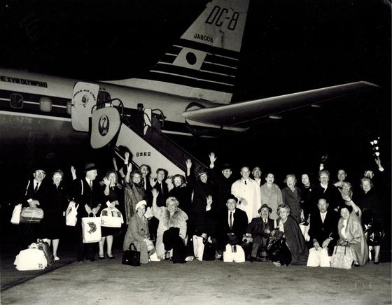 A tour group waving beside a Japan Air Lines DC-8. The plane carries the words "The XVIII Olympiad."