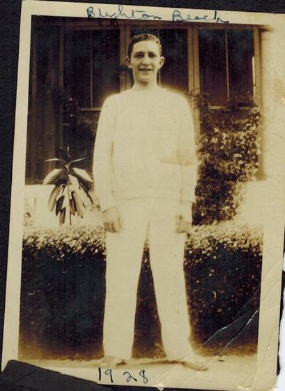 Harold, a smiling young man in a white sweater and white trousers in front of a hedge and a window.