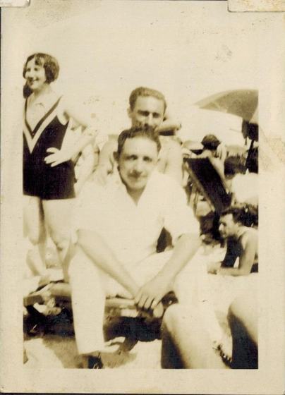 Harold in white, sitting in front on the sand, with a woman in a V-neck swimsuit and another man behind him.