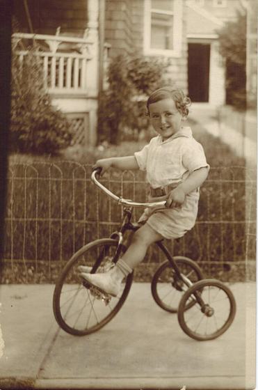 Harold as a little boy, riding a tricycle on the sidewalk in front of a wire fence and a porch.