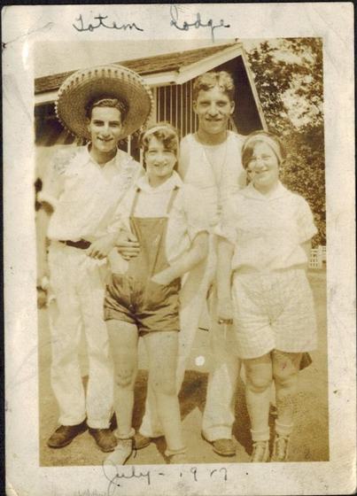 Four friends in front of a lodge: a young man in a big sombrero, a girl in short overalls, a tall man in a vest, and a girl in a headband and shorts.