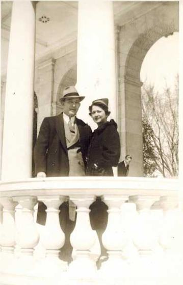 Harold and Mae between white columns above a stone balustrade, with bare trees behind them. He wears a fedora and overcoat, she a small hat and dark coat.