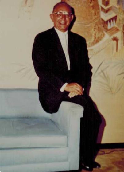 Harold sitting on the arm of a pale blue sofa in front of a painted mural, in a dark suit and glasses, smiling.
