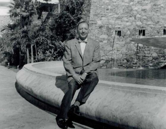 Harold in a sport jacket and bow tie, sitting on the edge of a stone fountain, with palm trees and a stone wall behind him.