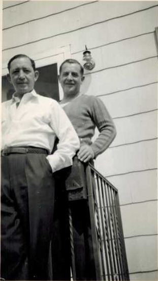 Harold and Tootsie at a railing below a shingled wall with a porch light: one in a white shirt with a hand in his pocket, the other in a sweater, smiling.