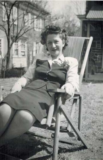 Mae in a vest, skirt and white blouse with a corsage, reclining in a wooden lawn chair.