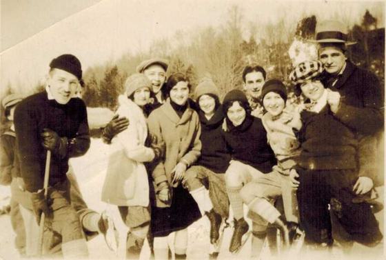 The Schnuer family on the snow: young people in winter coats and hats, some holding ice skates. One wears a checked cap and one a tall feathered hat.