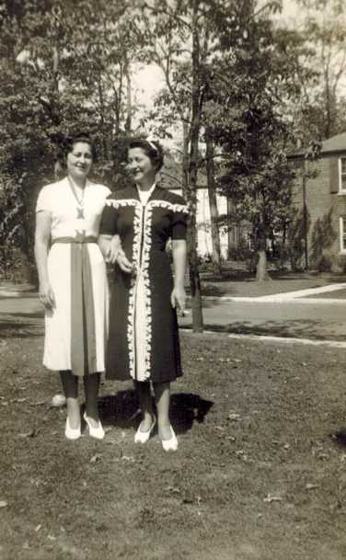 Mae with another woman, arm in arm on a lawn: one in a white dress with a belt, one in a dark dress with white trim, a brick house behind them.