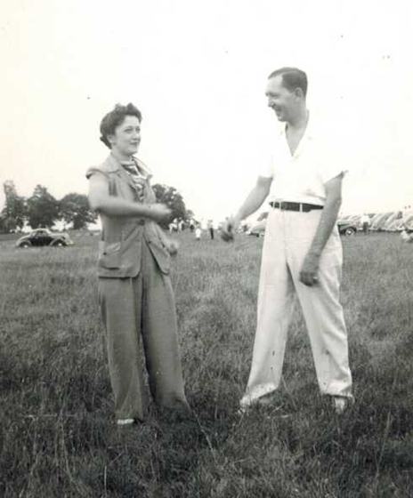 Harold and Mae talking on a field, with parked cars in the background.