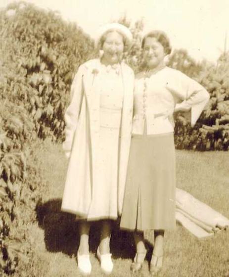 Mae and her mother-in-law, arm in arm in front of a hedge: one in a white coat and hat, one in a cardigan and pleated skirt.