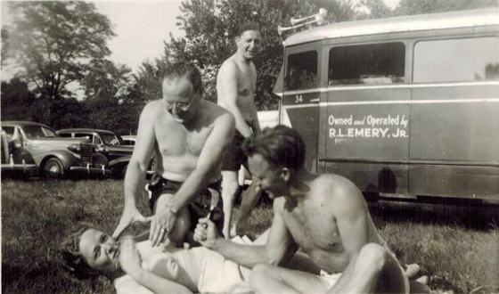 Harold and Tootsie, with others, on the grass beside a bus lettered "Owned and Operated by R. L. Emery, Jr." Three men are roughhousing and a woman lies on the ground laughing.