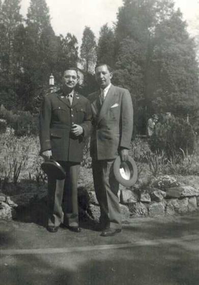 Harold with another man, both holding their hats: one in a military-style uniform, one in a suit, in front of a rock garden.