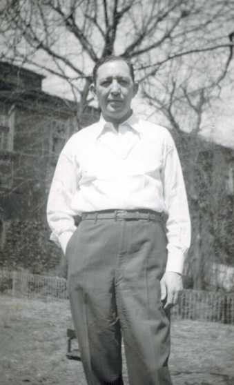 Harold in a white shirt and gray trousers, a hand in his pocket, outdoors beside a brick house.