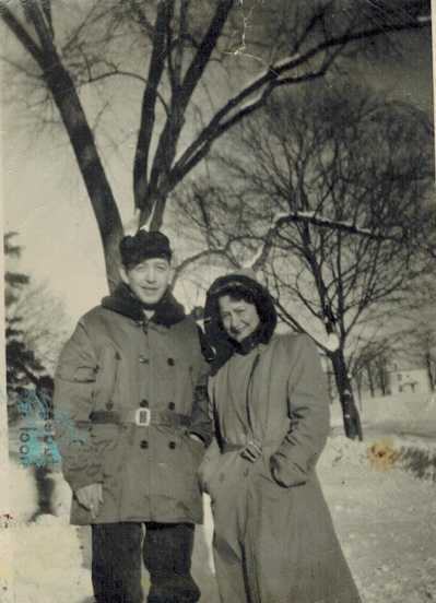 Harold and Mae standing in the snow beside a bare tree, he in a fur-collared coat with a belt, she in a long coat and hood.