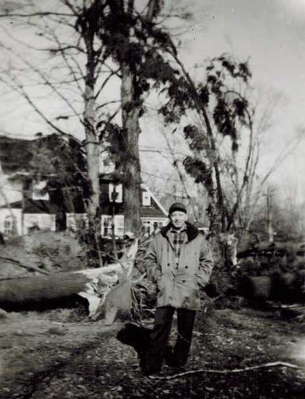 Harold in a hooded coat and cap, standing among fallen trees with a house behind him.