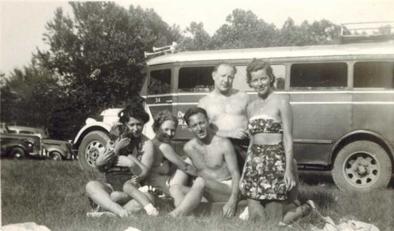 Harold and Tootsie with friends and a small child, sitting and kneeling on the grass in front of bus number 34, with old cars parked behind. The lettering starts "Ov... R..." (the same bus as P115 and P123).